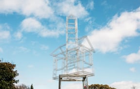 A sliver metal sculpture of a house frame on stilts.