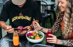 Two customers are eating buddha bowls and a glass bottle of juice is on the table.