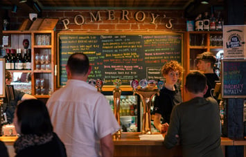 People queuing to order at the bar where on the back wall a big chalkboard lists all of the beers on tap in colourful chalk handwriting.