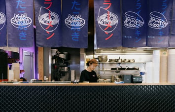 Worker standing behind counter and under Japanese style flags at Ramen Ria in Christchurch