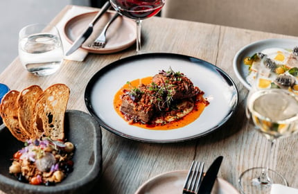 A meat dish on the table, surrounded by two other savoury dishes, glasses of wine and clean side plates.