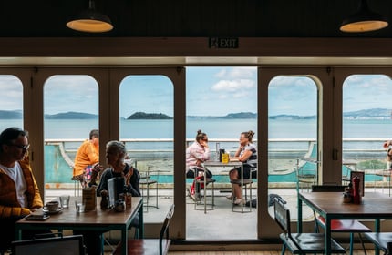 View from inside the first floor Seashore Cabaret cafe out to their deck area where people are dining with a view across the harbour to Wellington city beyond.