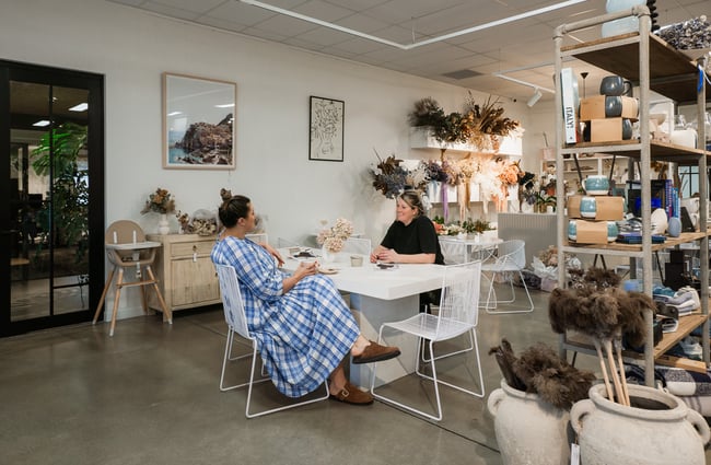 Two women sitting on white chairs inside a cafe and retail space.