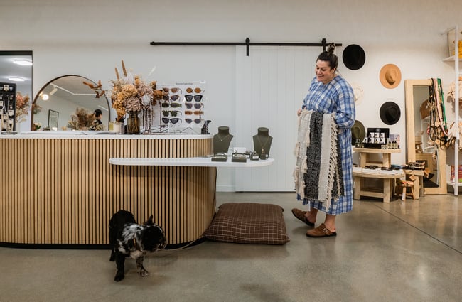 A woman wearing a blue and white dress smiling at a small black dog inside a retail store.