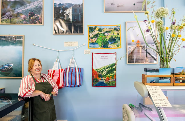 Julia standing in front of wall full of photographs, tea towels, and market bags at Akaroa Butchery