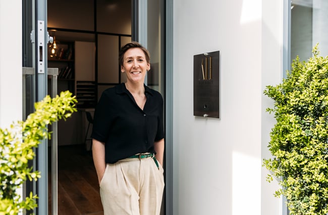 A woman with her hands in her pockets standing outside her jewellery store, smiling to camera.