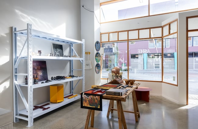 Large window bringing light into the gallery space, shelves and tables with art displays at Ashe.