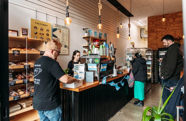 Customers standing around counter ordering and staff making coffees at Aurora Argentinian Bakery.