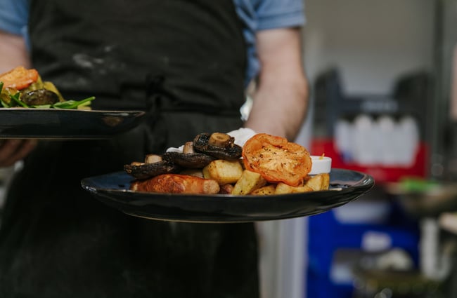 A waiter carrying a black plate with a sausage, tomato, egg and potato breakfast meal.