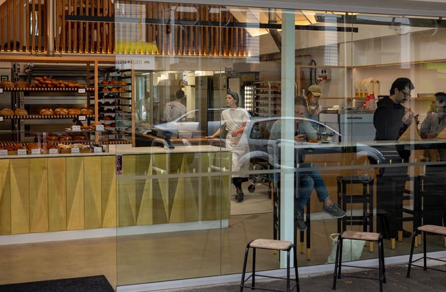 Looking into the bakery through the glass frontage; bread on the shelves and customers sitting at the leaner in the window.