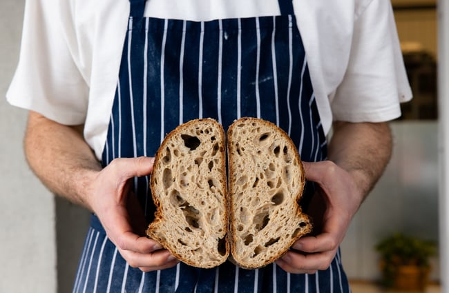 A man wearing a blue and white apron holding a loaf of bread cut in half.