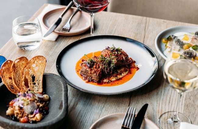 A meat dish on the table, surrounded by two other savoury dishes, glasses of wine and clean side plates.