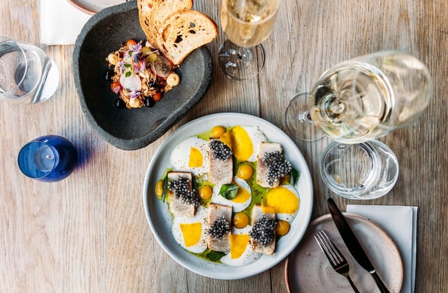 Birdseye view of a table laid with glasses of wine, a plate of pieces of fish with multiple toppings and garnishes, a small candle in a blue glass and an asymmetric dish of toast and bits.