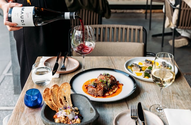 A waiter pours red wine into a large wineglass on a table laid with three savoury dishes and two table settings.