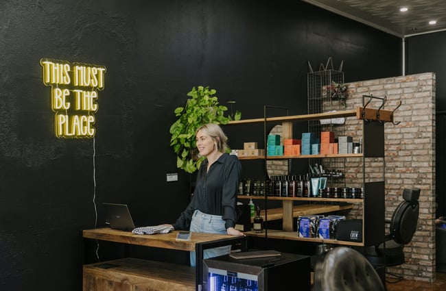 A blonde haired woman smiling behind the counter at Bruce's Barber Oamaru. '