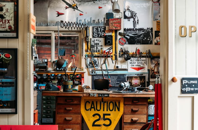Workshop space filled with tools and scrap materials with a big yellow 'caution' sign at the front of the desk