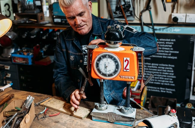Close up of Martin using a tool to work on orange robot sculpture at his workshop desk