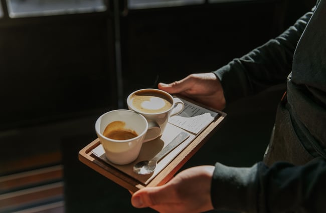 Close up of a staff member carrying a small wooden tray with a black and a white espresso coffee on it.