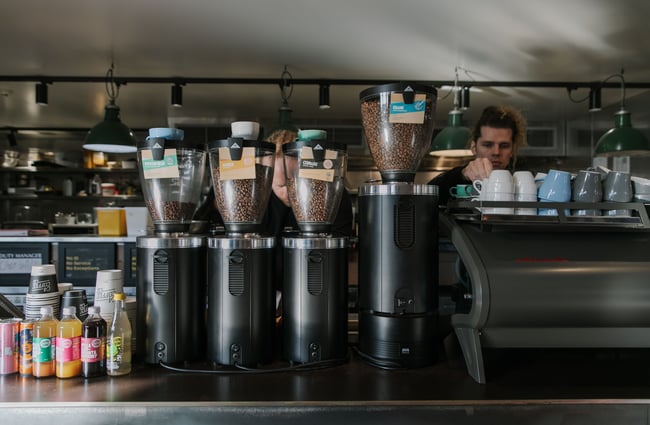 Two baristas working behind the coffee machine and four coffee bean grinders housing four different types of beans.