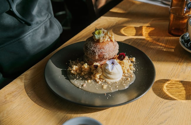 Close up of deep-fried pancakes coated in sugar, topped with pickled green apple and served with caramel mascarpone and maple butter, garnished with crumbles and pansy flowers.