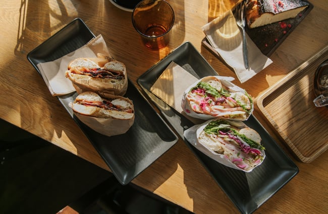 Birdseye view of a wooden table top at C4 with two separately plated bagels and a slice of cheesecake, casting shadows in the sun.