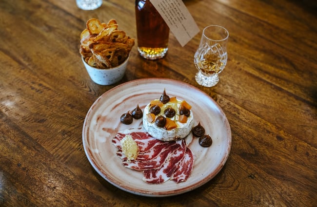 A plate of baked camembert, little bowl of crostini and dram of whisky on a wooden table at Cardrona Distillery.