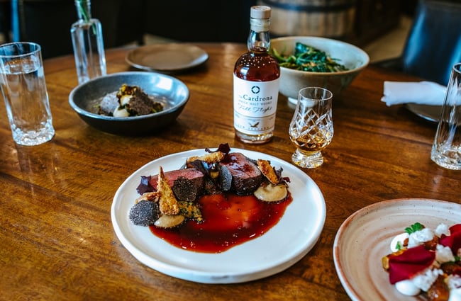 A venison dish, other savoury plates and a dram of whisky spread across a wooden table at Cardrona Distillery.