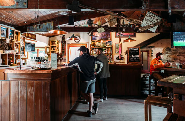A few people ordering at the bar at Cardrona Hotel, low wooden trusses overhead are decorated with old mining paraphernalia.