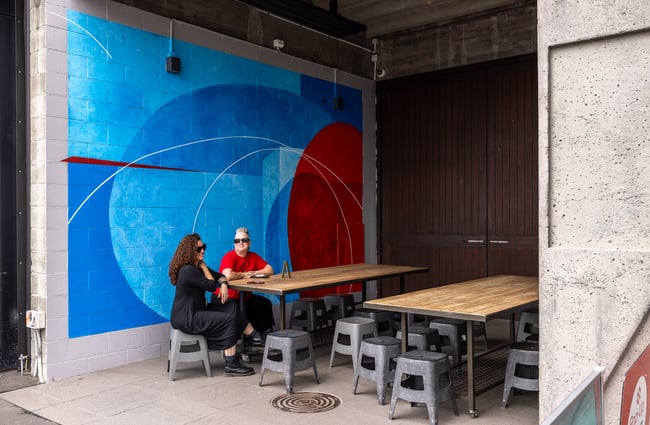 Two people sitting at table in front of bright mural at CoCA Gallery in Christchurch