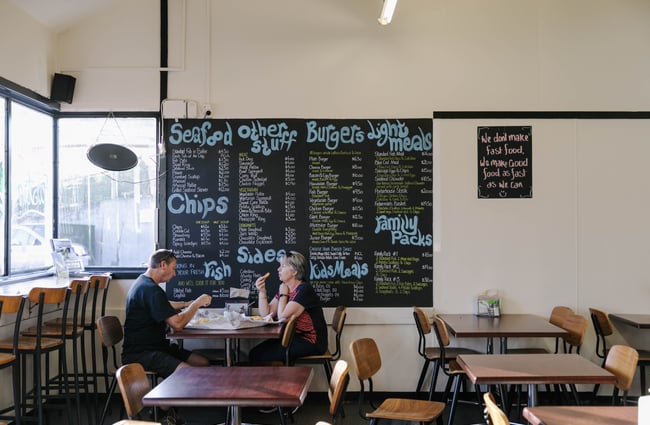 Couple eating fish and chips at a table in front of menu board.
