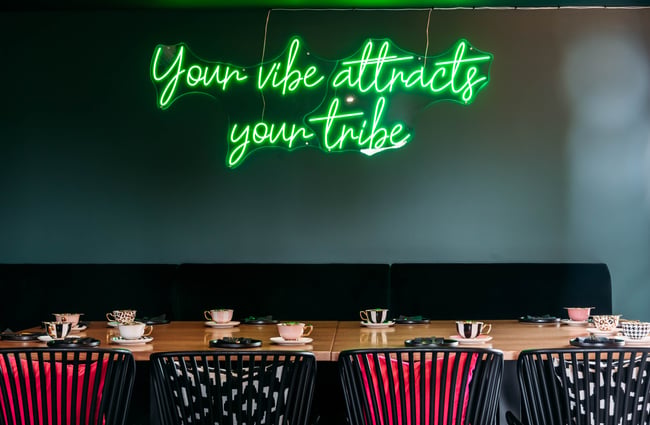 Above a table and chairs setting on the wall is a lit up sign that reads 'Your Vibe Attracts Your Tribe' at Country Road High Tea.