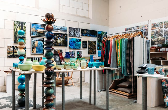 Interior of Crafted Gallery in Ōamaru with a table filled with ceramics, photography on the wall and fabrics hanging from metal rack