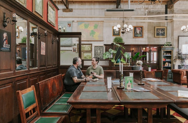 Two people sitting in the far corner of a big square wooden table inside Craftwork Brewery.