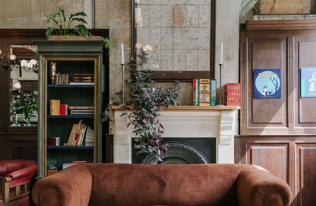 A brown suede couch in front of an old fire place decorated with books and plants.