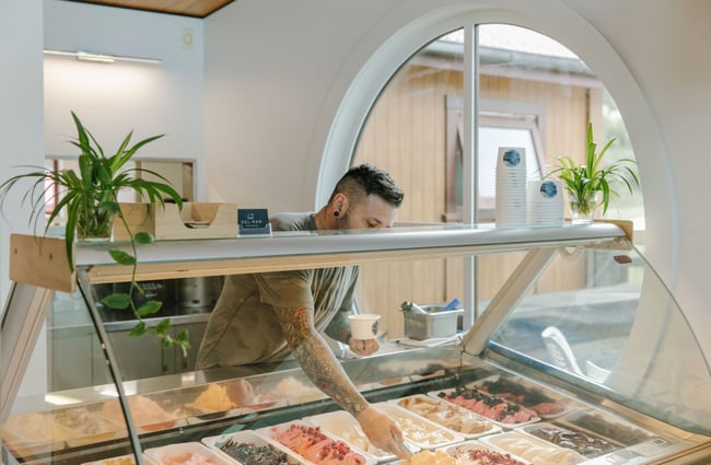 A man in a grey t-shirt scooping gelato into a tub for a customer.