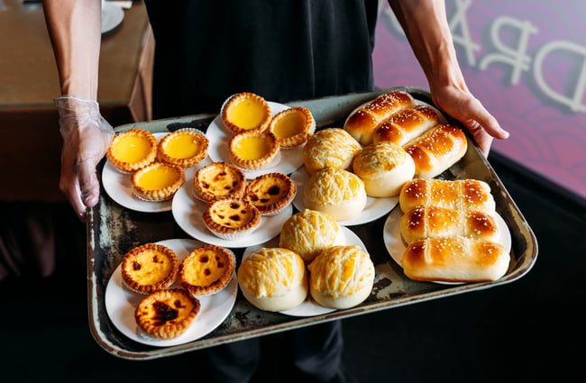 A person holding a large tray of plates of various custard egg tarts and soft white bread buns.