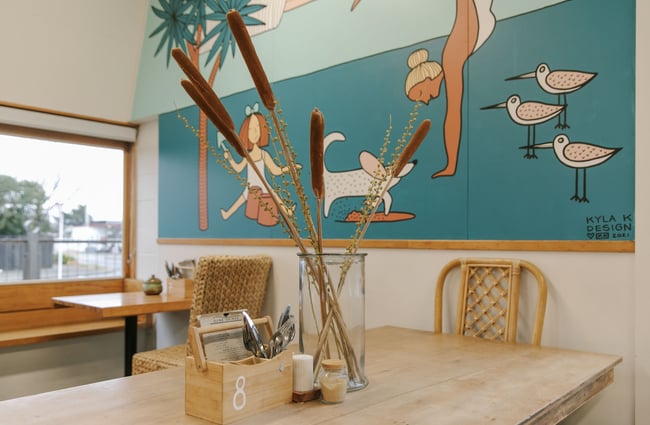Close up of a table setting with a large glass jar of dried reeds, and a blue-brown painted beach mural on the wall behind