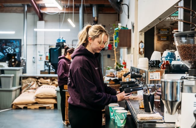 Barista behind machine, with coffee bean roasting area behind at Ebony Coffee Roasters.
