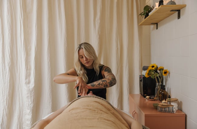 Front on view of beauty therapist performing facial massage with a vase of sunflowers on cabinet next to bed