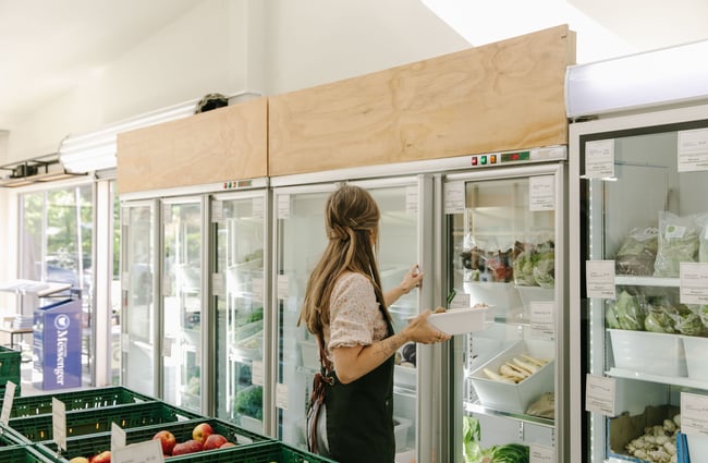A lady in a black Freshlink Grocer apron holding a white tub about to put it into the fridge.