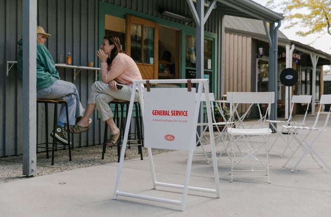 A sign showing the shared work space with two people having coffee at a bench.