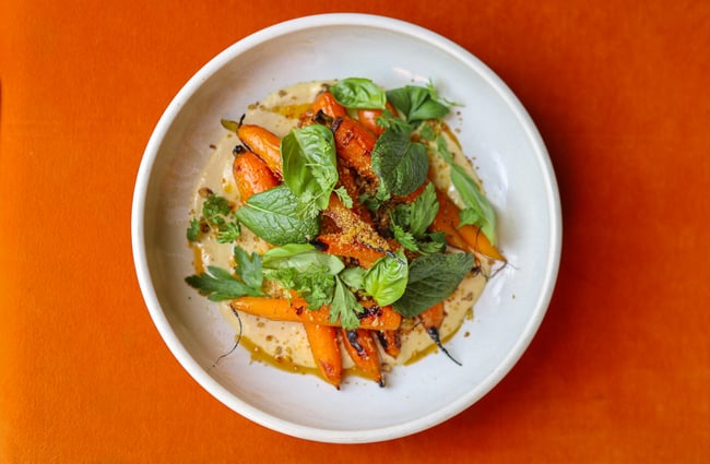 Plate of carrots on hummus with green herb leaves, on a white plate on a bright orange table at gin gin.