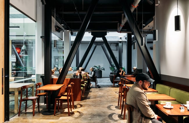 Interior of Glou Glou with people dining at wooden tables and green suede booths with black industrial beams running across the ceiling