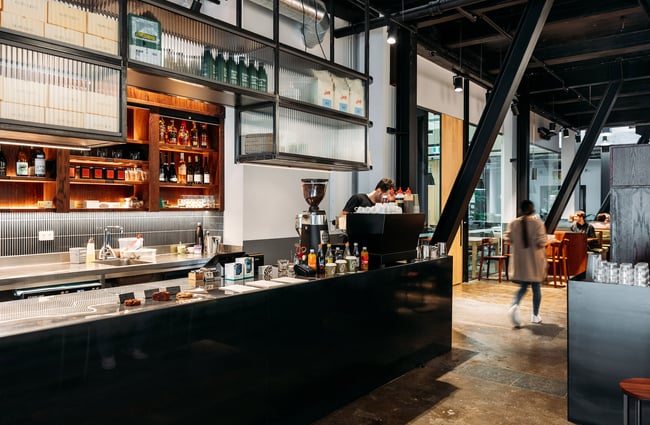 Male barista standing behind black counter of Glou Glou in Wellington with rippled glass cabinets above and baked goods presented in a clean and simple cabinet
