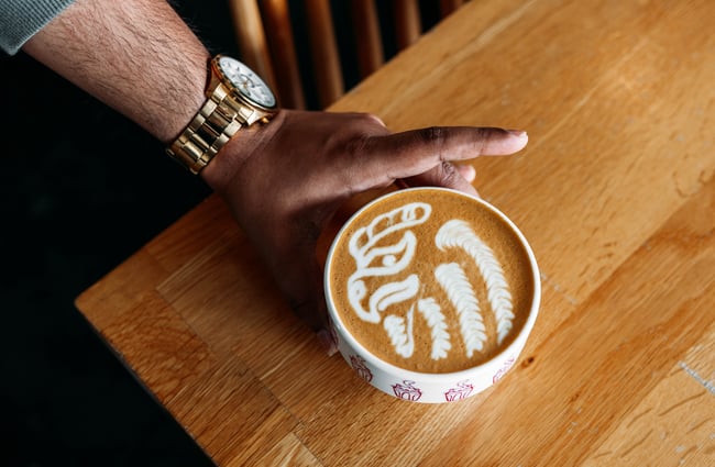 Close up of an arm wearing a big gold watch placing a mug of coffee on a wooden table on the top of which a detailed phoenix has been drawn with steamed milk.