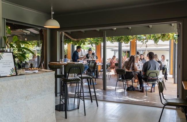 View from the interior dining room of people dining out on the verandah, covered by a canopy of leafy vines, at Greystone Wines, North Canterbury.