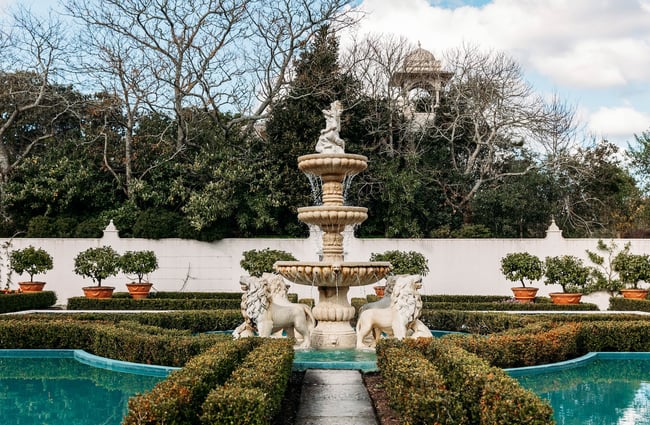 A water fountain surrounded by marble lion statues and symmetrical low hedging.