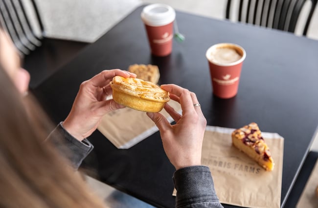Customer holding a pie, on the table in the background there is a coffee and a slice at Harris Farms Butchery, Bakery & Café.