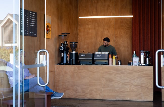 Looking through glass door entrance of cafe with a barista standing behind coffee machine and customer sitting waiting