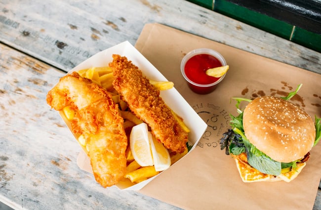 A flatlay of fish and chips and burger with a little pottle of sauce on an outdoor table.